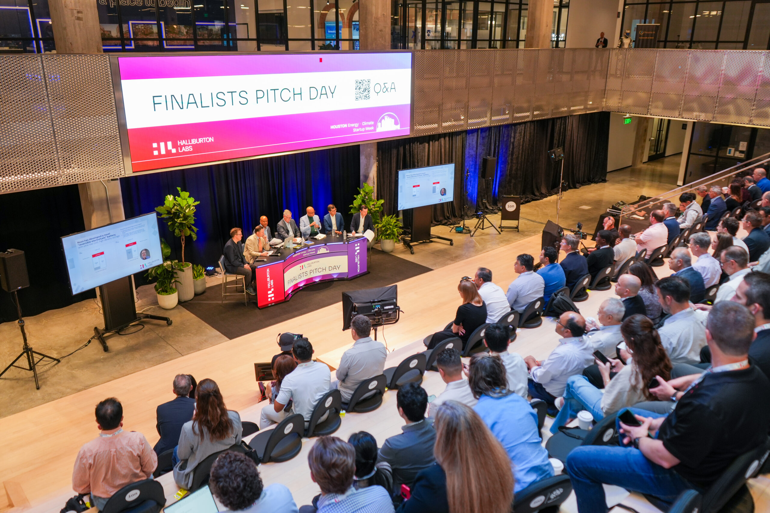 A panel of five men sits at a curved stage desk during a 'Finalists Pitch Day' Q&A session at Halliburton Labs, part of Houston Energy & Climate Startup Week, held inside The Ion. A large overhead screen displays the event title, and a seated audience faces the stage, with presentation slides visible on a side monitor.